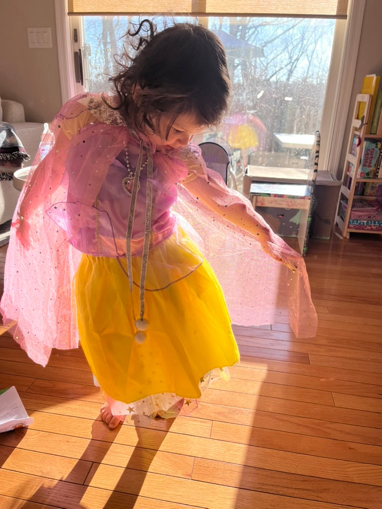 Child in a pink and yellow dress standing in a room with wooden flooring.
