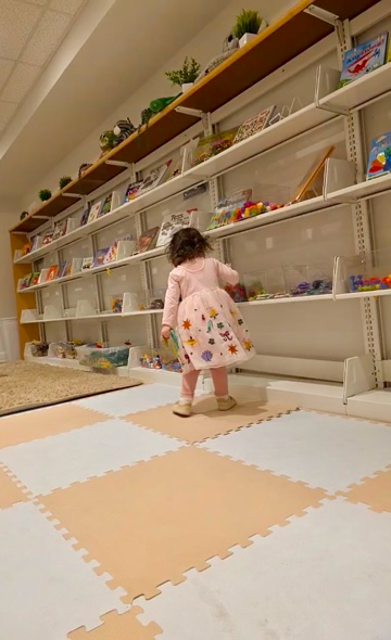 Child in a pink dress standing on a foam play mat in a library with shelves and toys